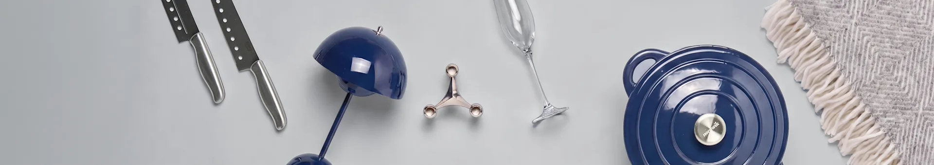 Surtido de menaje de cocina y decoración del hogar: dos cuchillos plateados, una lámpara decorativa azul, un utensilio de cocina plateado, una copa de champán, una olla azul con tapa y una manta estampada.