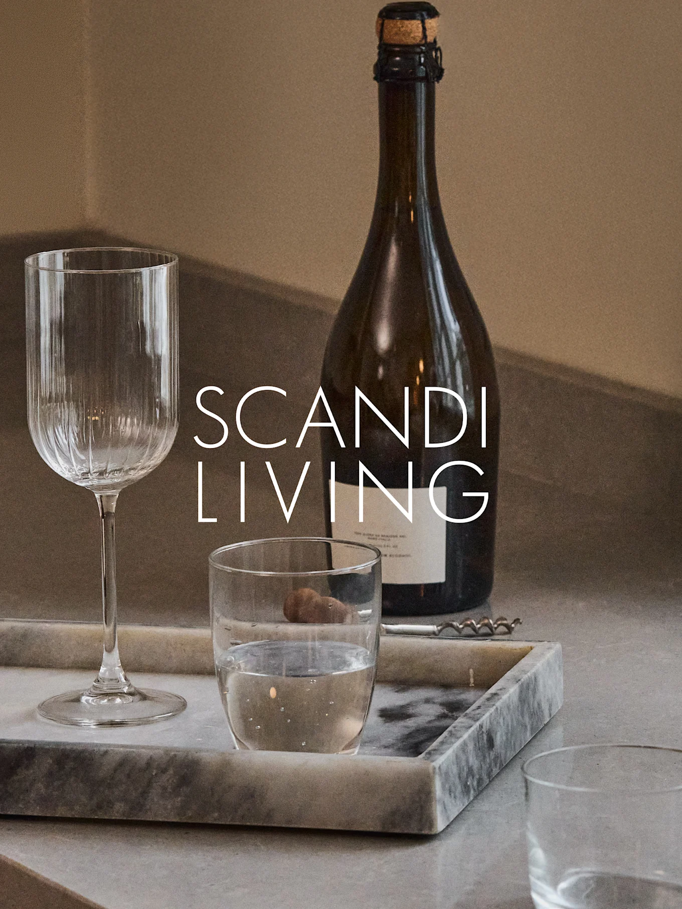 Una botella oscura, una copa de vino acanalada, un vaso de agua y un sacacorchos en una bandeja de mármol con el texto 'SCANDI LIVING'.