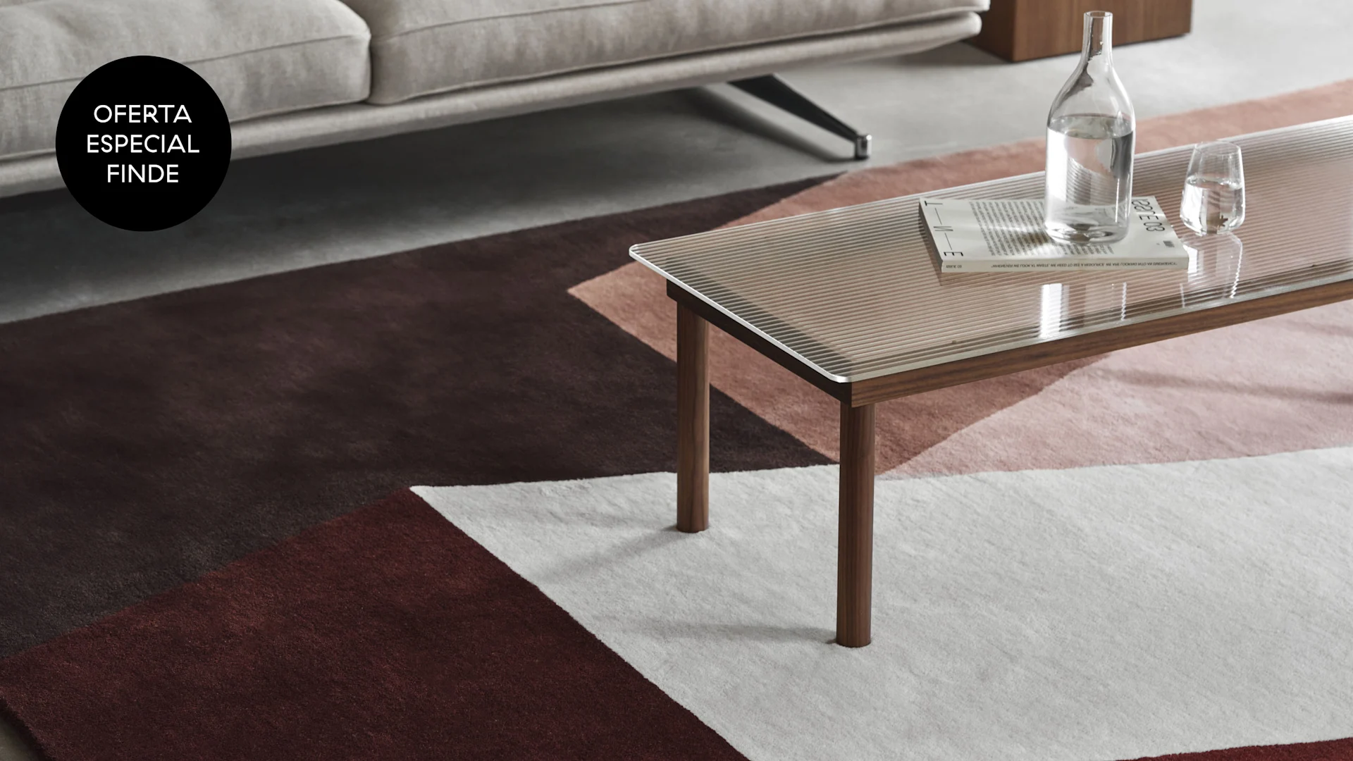 Sofá, mesa de centro de cristal y madera, y alfombra geométrica marrón, rosa y blanca. Etiqueta negra: 'Oferta Especial Finde'.