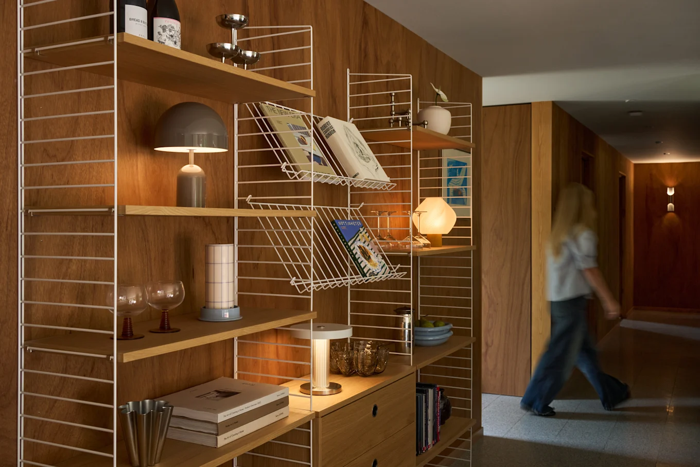 Una persona rubia camina por un pasillo de madera junto a una estantería moderna llena de libros, lámparas y objetos decorativos.
