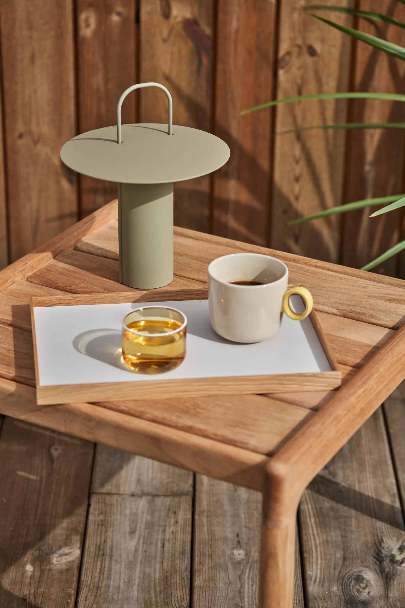 Una lámpara portátil verde claro, un vaso con líquido ámbar y una taza de café color crema en una bandeja de madera sobre una mesa de madera exterior.