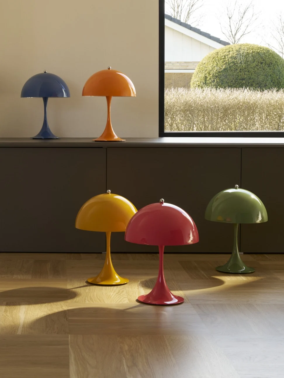 Seis lámparas coloridas con forma de seta, dos sobre un mueble y cuatro en un suelo de madera, junto a una ventana con vistas a un jardín.
