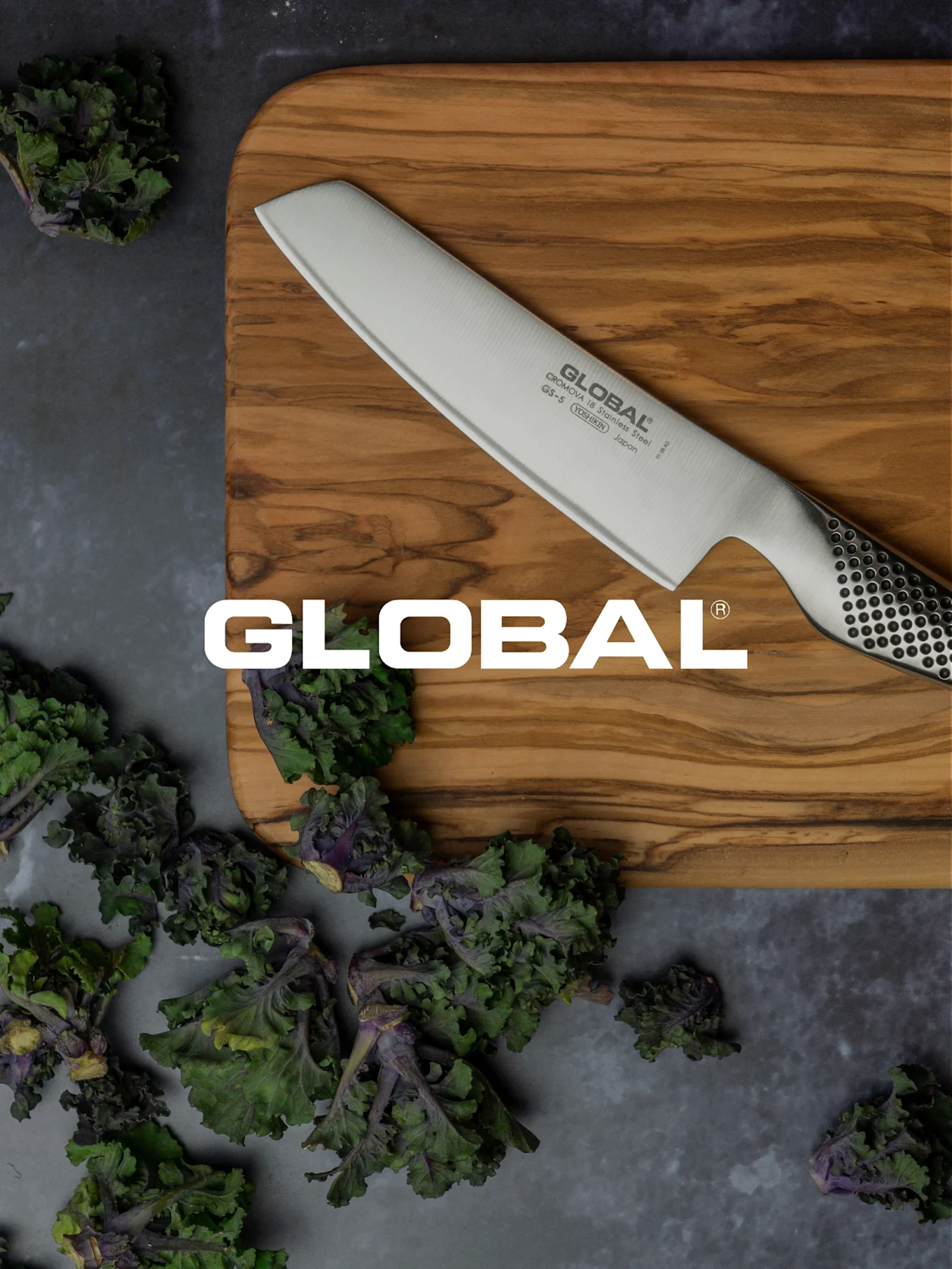 Un cuchillo Global sobre una tabla de cortar de madera, rodeado de verduras de hoja verde y moradas frescas.