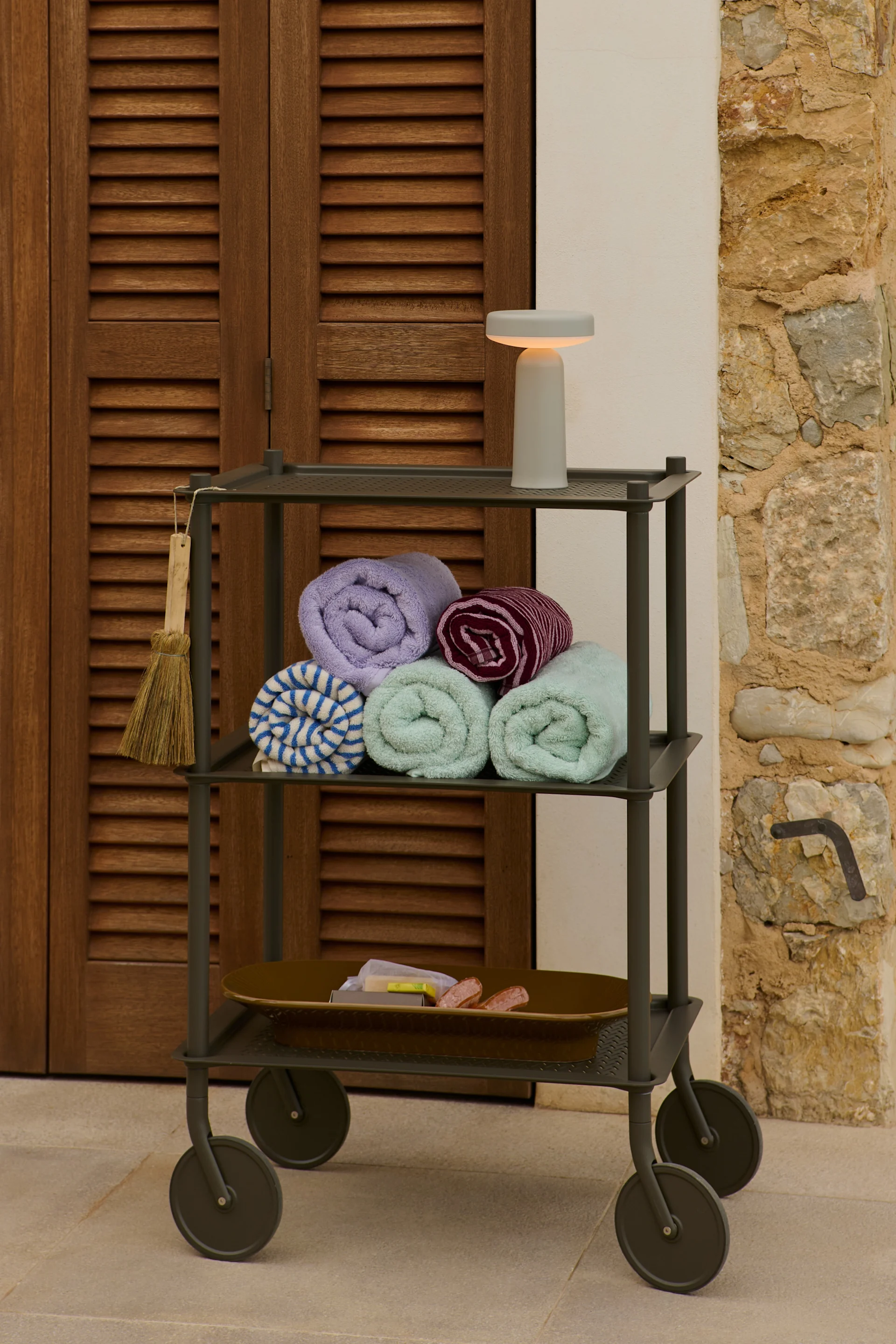 Un carrito utilitario de tres niveles con toallas enrolladas, artículos de tocador y una lámpara junto a una puerta de madera y una pared de piedra.