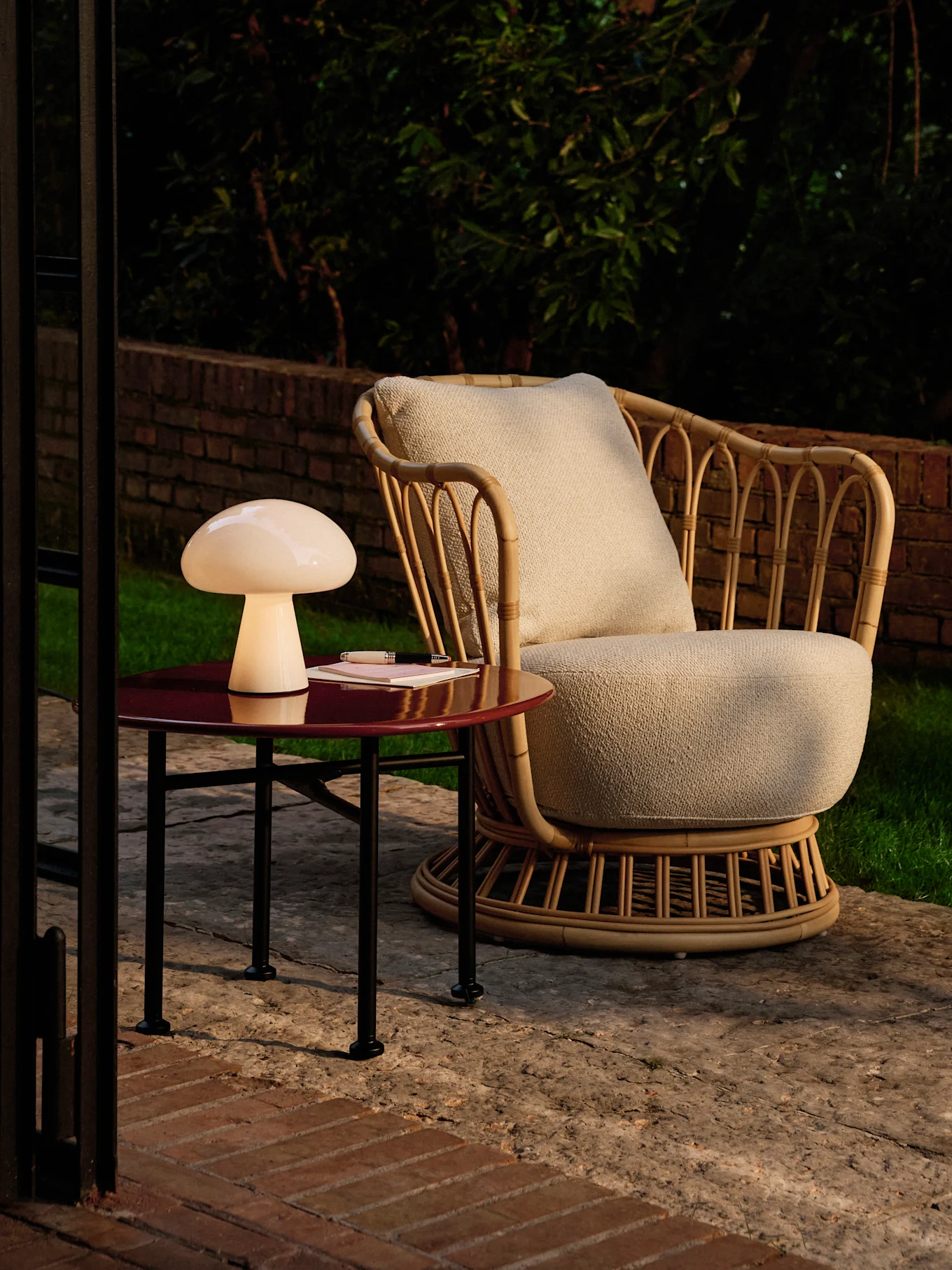 Lámpara seta en una mesa auxiliar roja junto a un sillón de ratán en un patio al anochecer.