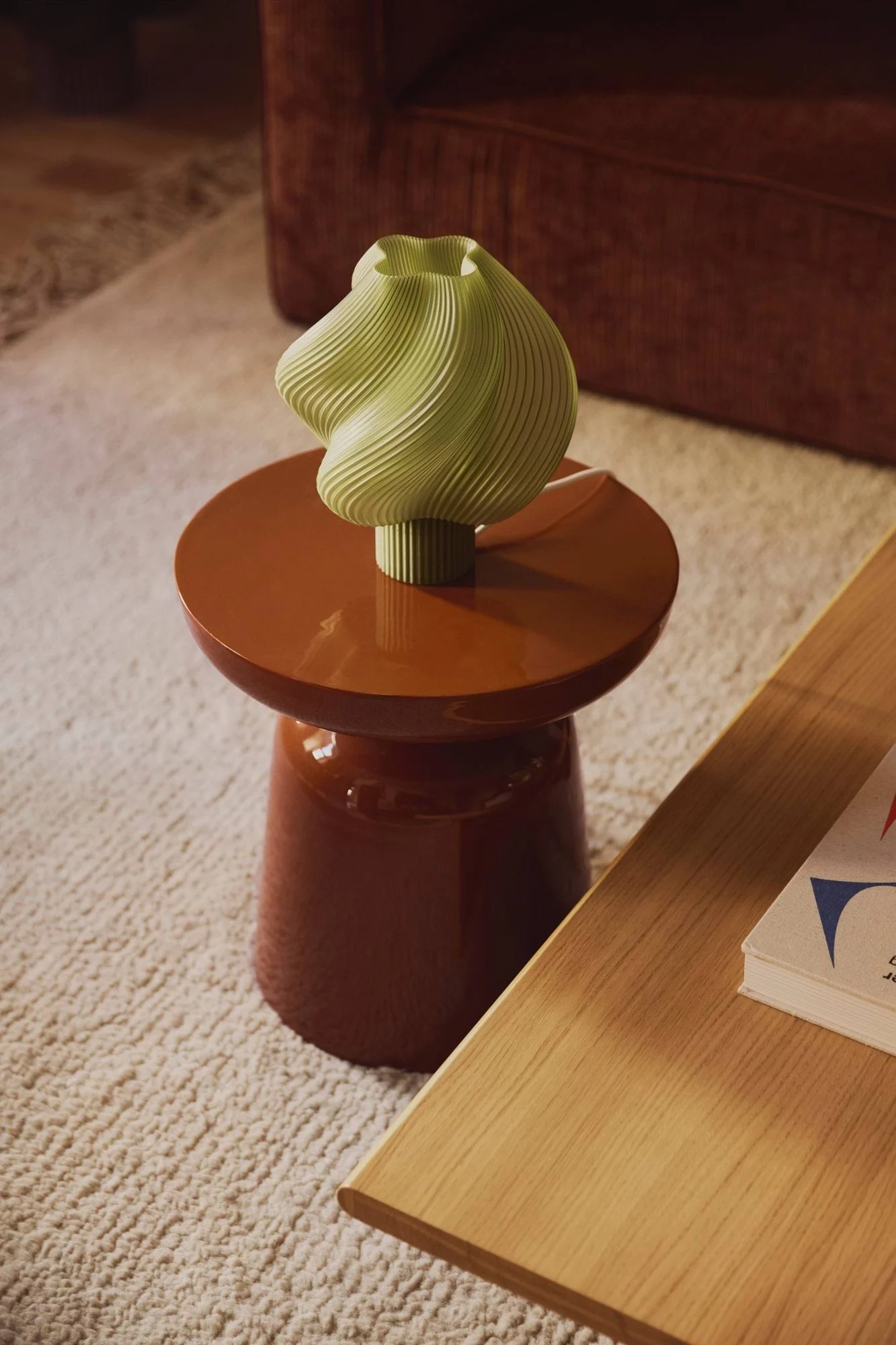 Un jarrón verde claro y estriado descansa sobre una mesa auxiliar redonda de color marrón rojizo, en una alfombra crema con un sofá al fondo.