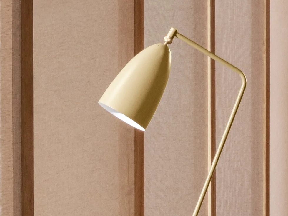Un primer plano de una lámpara moderna de color dorado amarillento con un cabezal cónico inclinado hacia abajo, que emite luz contra una pared de paneles marrón claro.
