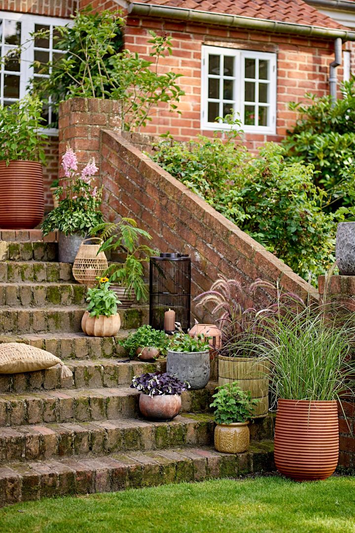 En una escalera del jardín hay varias macetas de estilo boho con plantas dentro, entre ellas la maceta Mesola de Bloomingville.