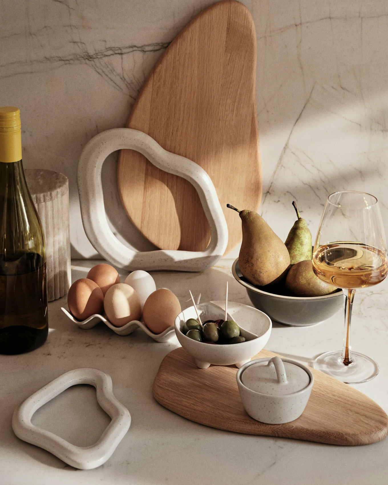 Encimera de mármol con botella de vino, huevos, aceitunas, peras y artículos decorativos de cerámica y madera.