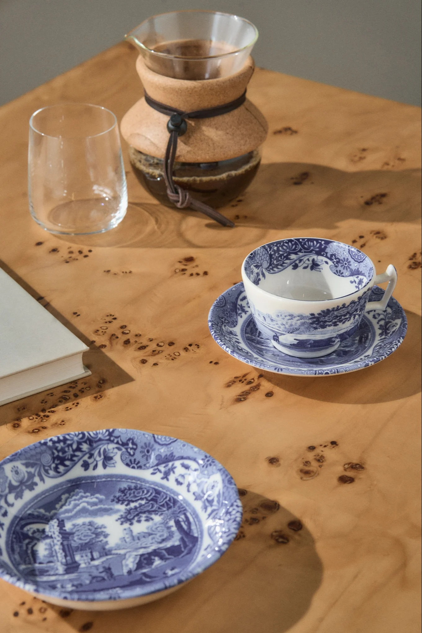 Cafetera de goteo, un vaso vacío, una taza de té azul y blanca con platillo, y un plato a juego sobre una mesa de madera clara.