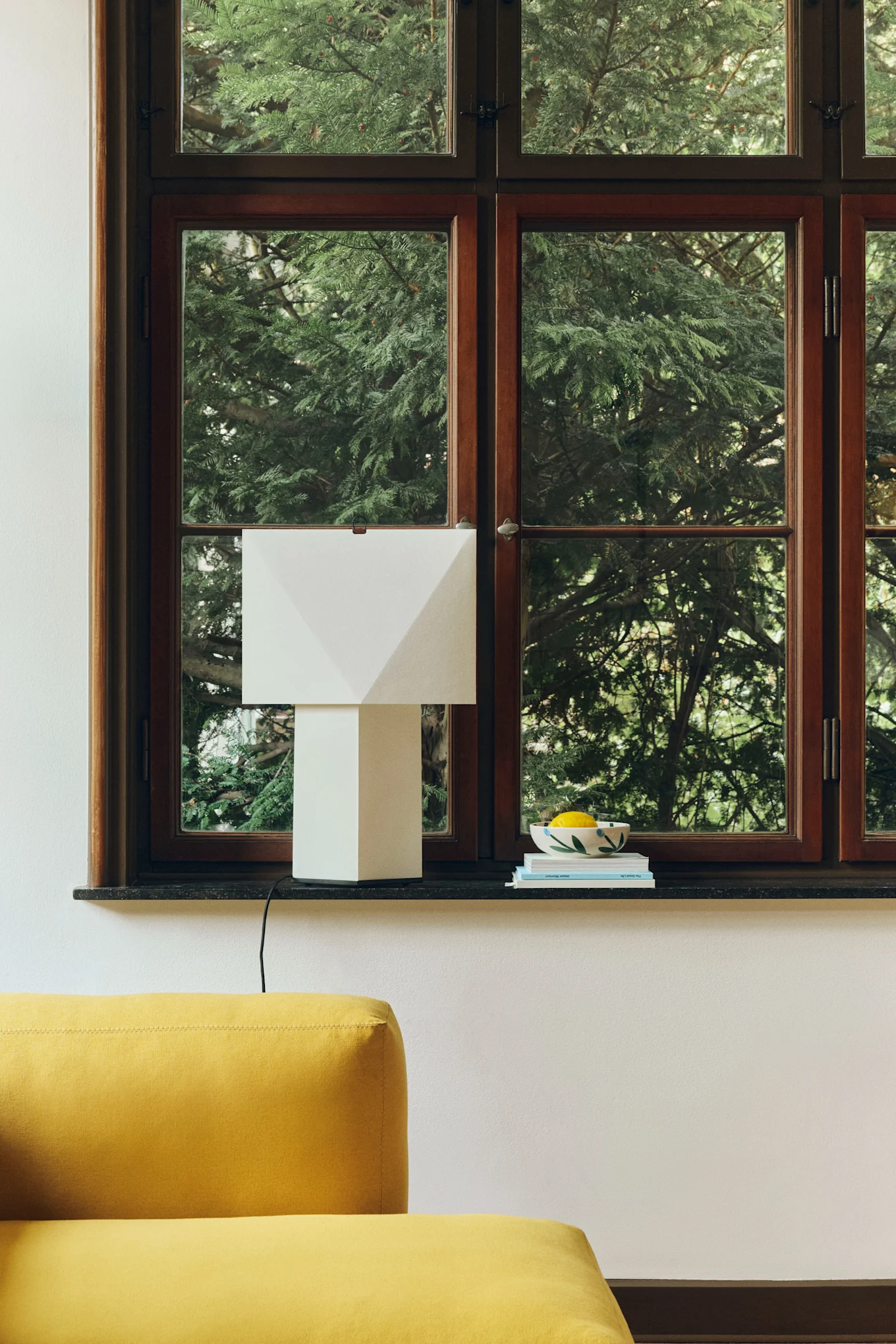 Una lámpara de mesa geométrica blanca sobre un alféizar oscuro junto a un cuenco de limones y libros, con un sofá amarillo en primer plano.