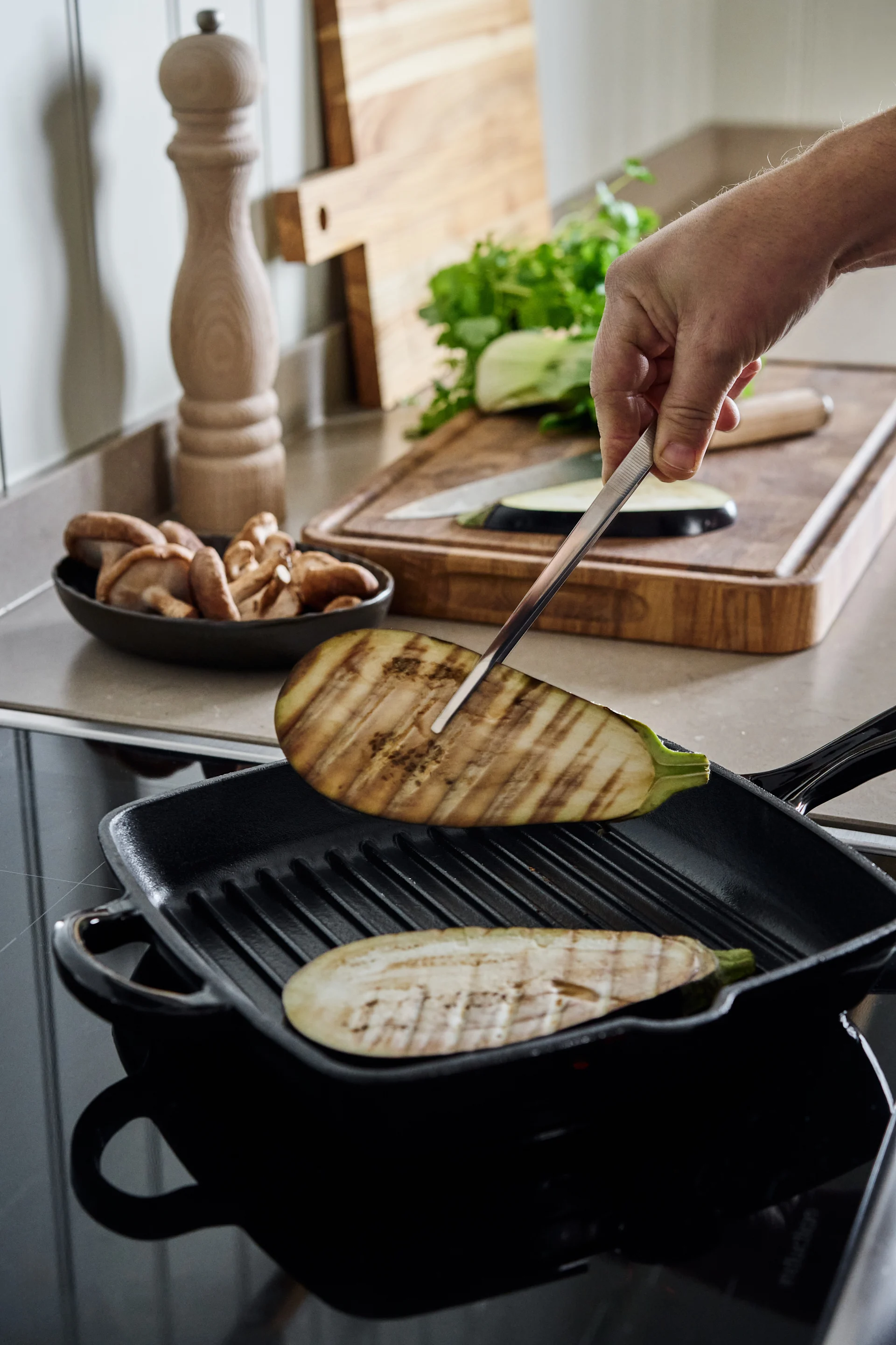 Mano volteando rodaja de berenjena a la parrilla en sartén negra sobre placa de inducción, con champiñones y verduras.