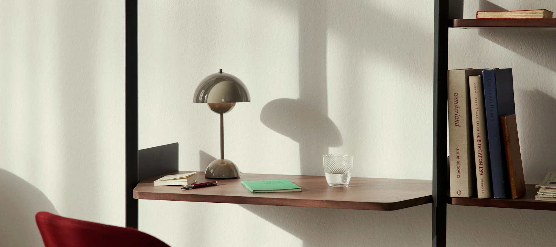 Lámpara de escritorio moderna con forma de hongo, un vaso de agua y cuadernos en un estante de madera junto a una librería.