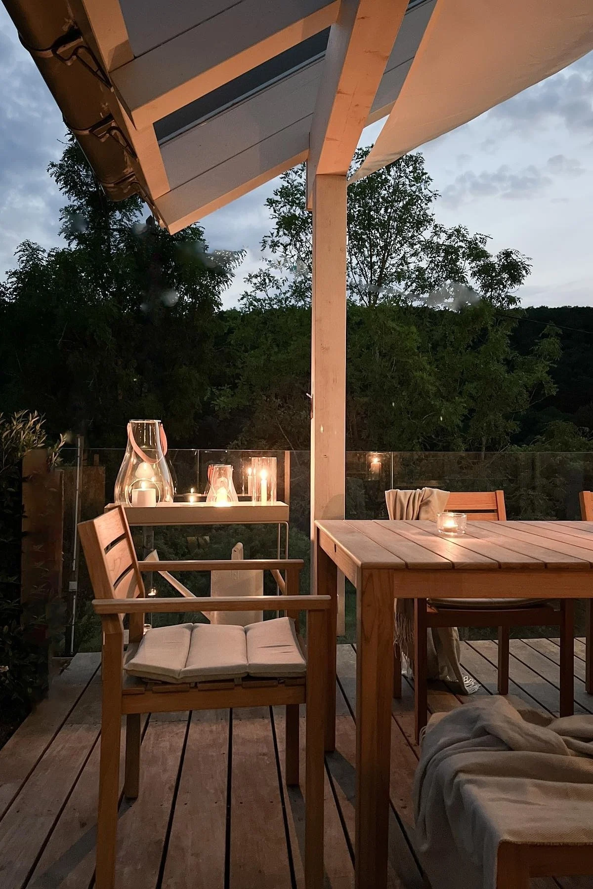 Coloca varios farolillos y portavelas en el exterior de tu patio para crear un ambiente acogedor. Aquí puedes ver el farol Design with Light de Holmegaard sobre la mesa de patio de madera de la influencer alemana @zuhauselig.