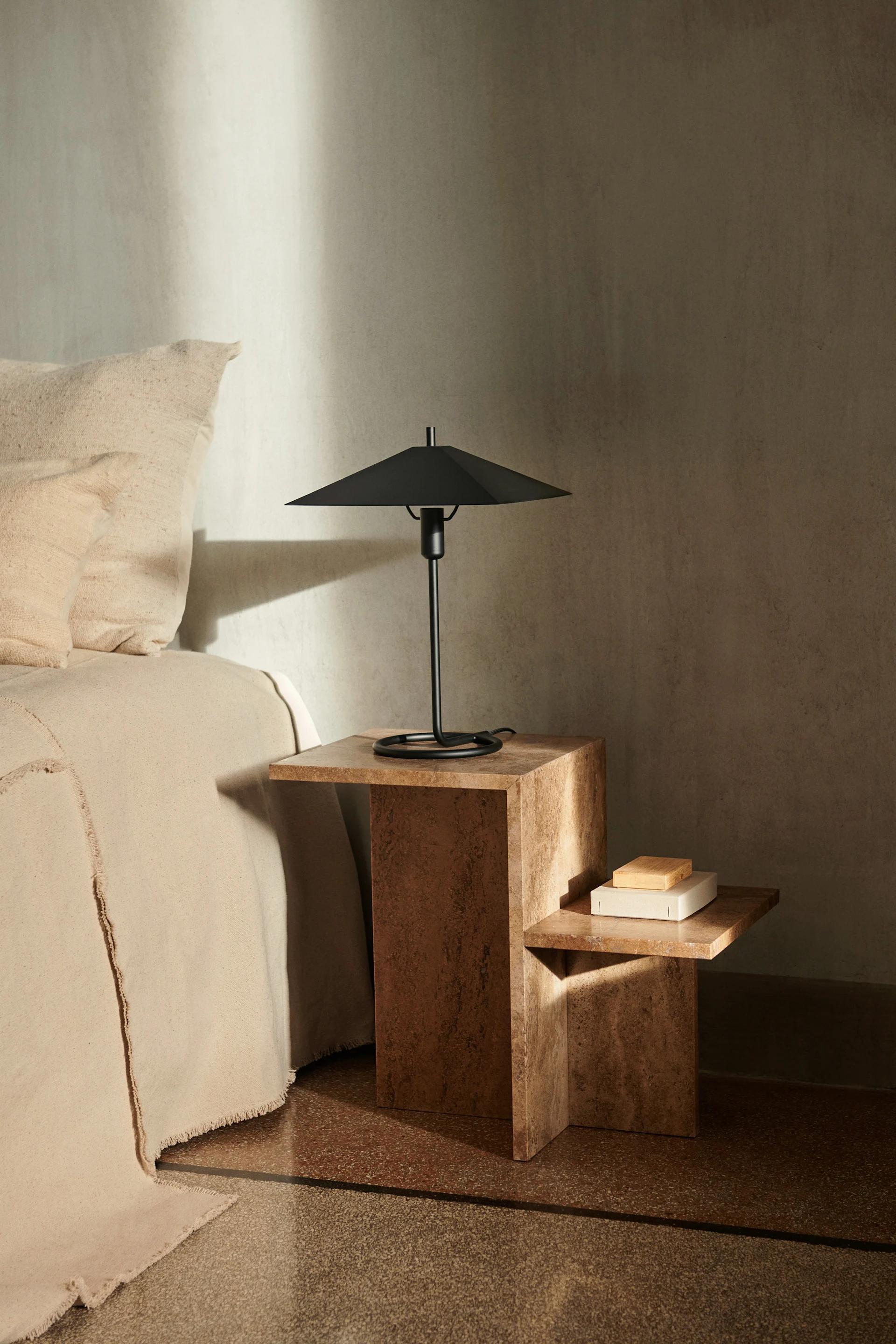 Escena de dormitorio con una cama beige junto a una mesita auxiliar marrón texturizada de dos niveles, con una lámpara de mesa negra moderna.