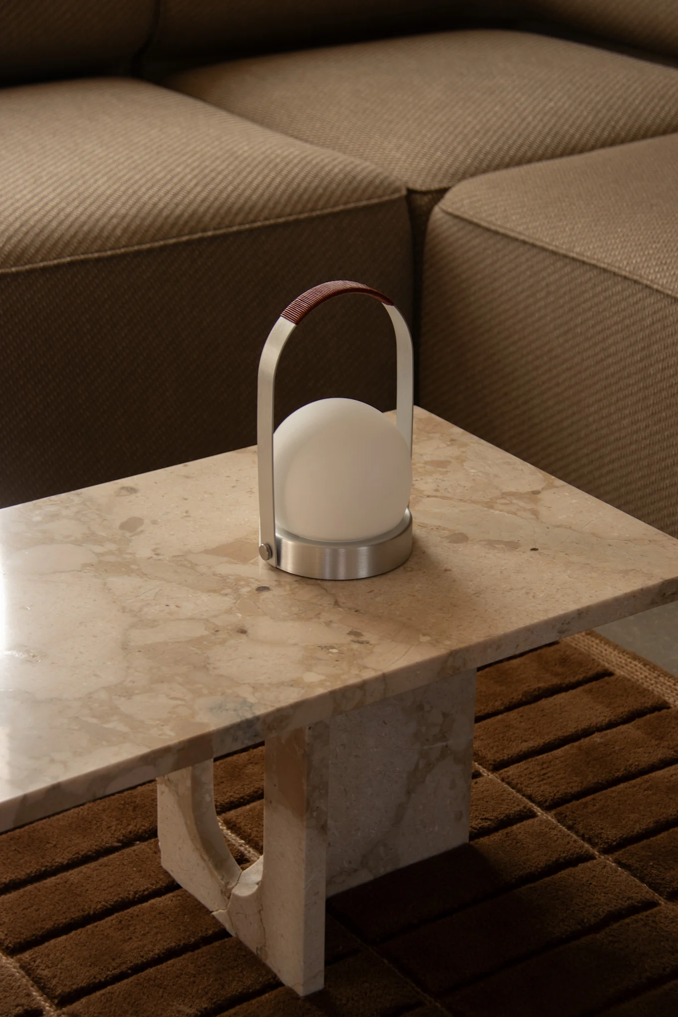 Una lámpara portátil moderna con luz esférica blanca, marco de metal cepillado y asa de cuero marrón sobre una mesa de mármol claro.