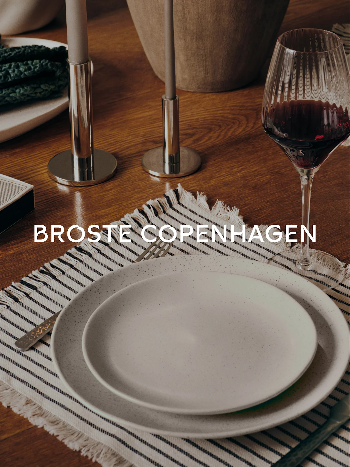 Mesa de comedor con dos platos moteados sobre un mantel individual a rayas, tenedor, copa de vino, dos candelabros y el texto 'BROSTE COPENHAGEN'.