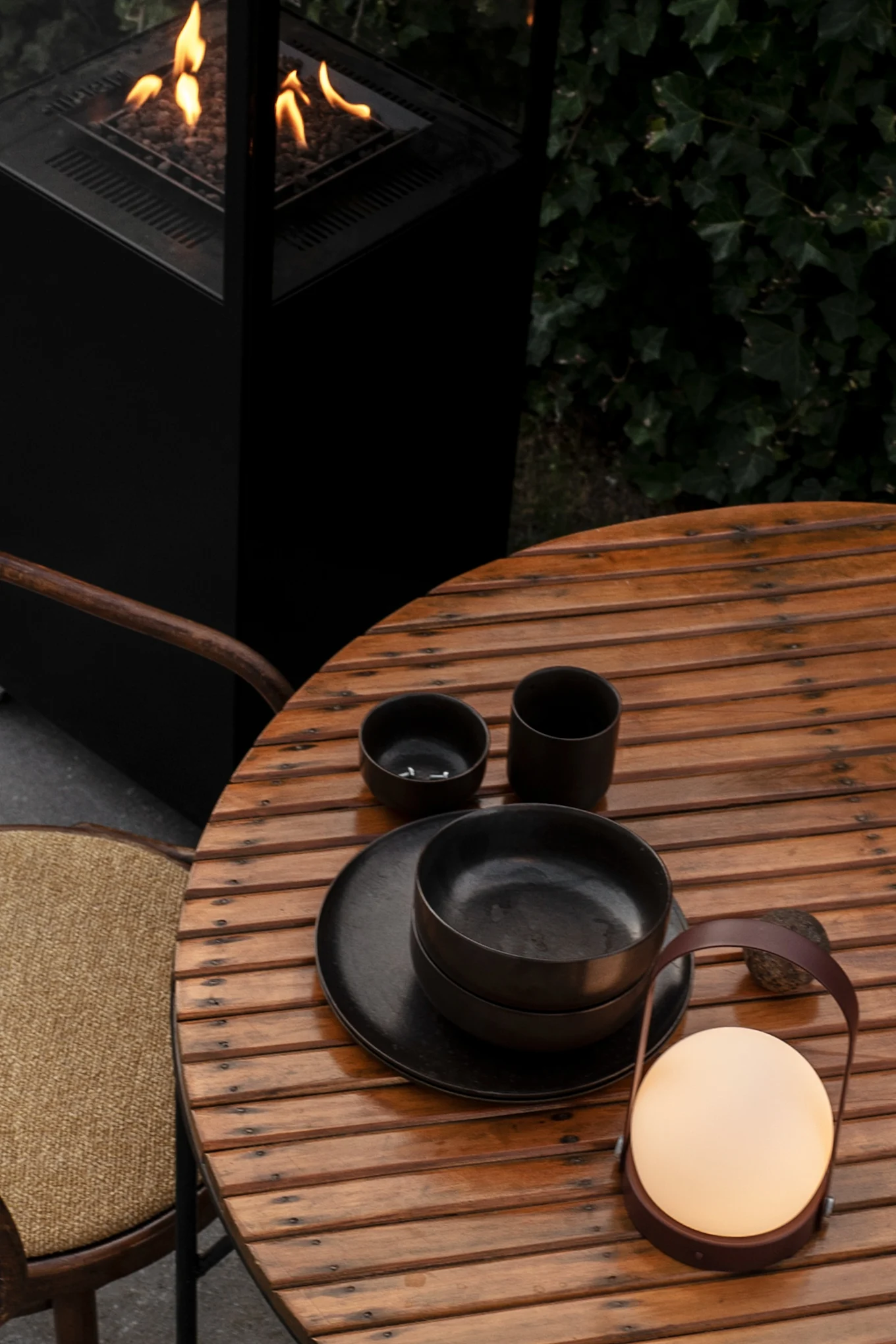 Aquí se puede ver la lámpara Carrie en Burned Red, también apta para exteriores, sobre una mesa de jardín de madera, junto a un servicio de mesa negro. A su lado hay una silla y, detrás de la mesa, arde un brasero.