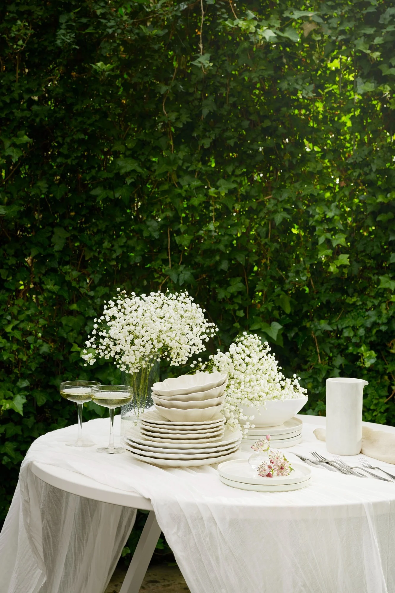 Mesa exterior puesta con vajilla blanca, flores de paniculata, copas de champán y cubiertos sobre un mantel blanco, contra hojas verdes exuberantes.