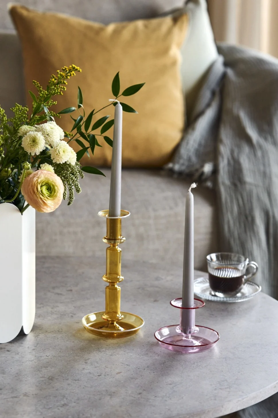 Dos candelabros de cristal, uno ámbar y otro rosa, con velas grises en una mesa junto a un jarrón de flores y una taza de café.
