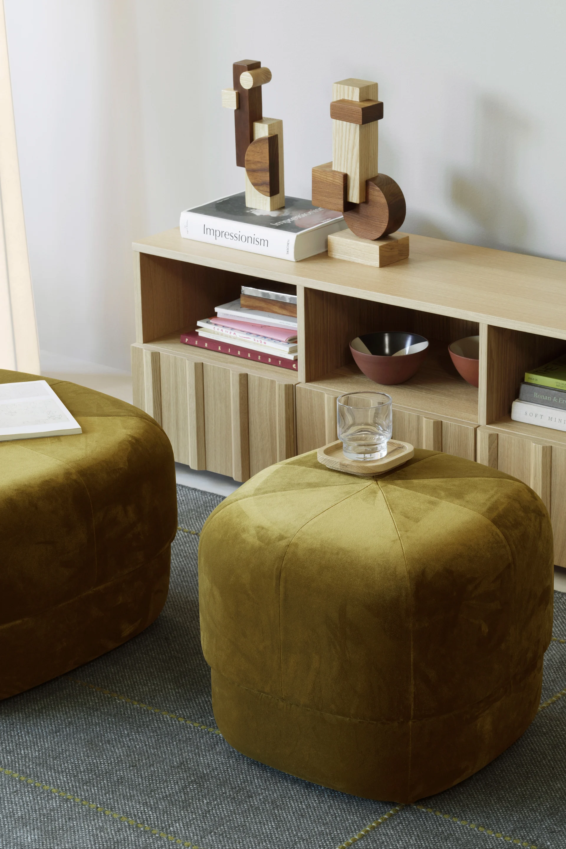 Dos otomanas de terciopelo verde oliva sobre una alfombra estampada junto a una estantería de madera clara con libros y decoración de madera.