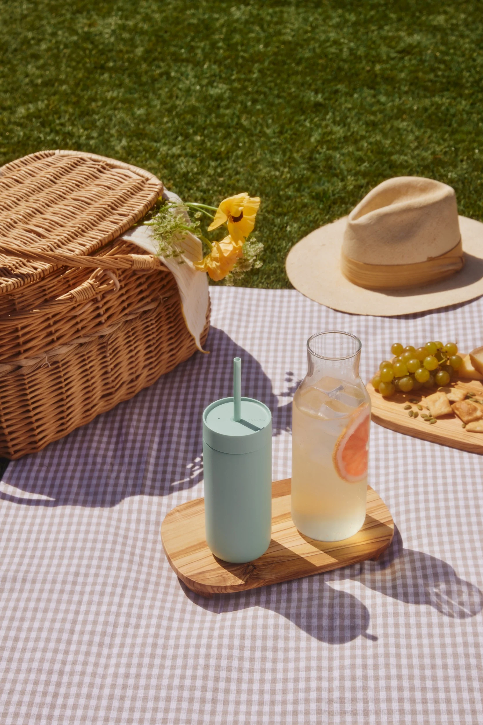 Picnic en una manta a cuadros: cesta de mimbre, vaso verde claro, bebida de pomelo, uvas y galletas saladas sobre la hierba.
