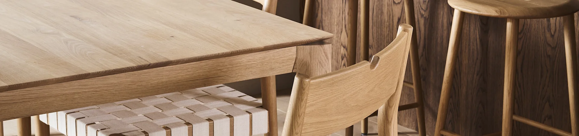 Primer plano de una mesa de comedor de madera clara con una silla de asiento trenzado y taburetes de madera al fondo.