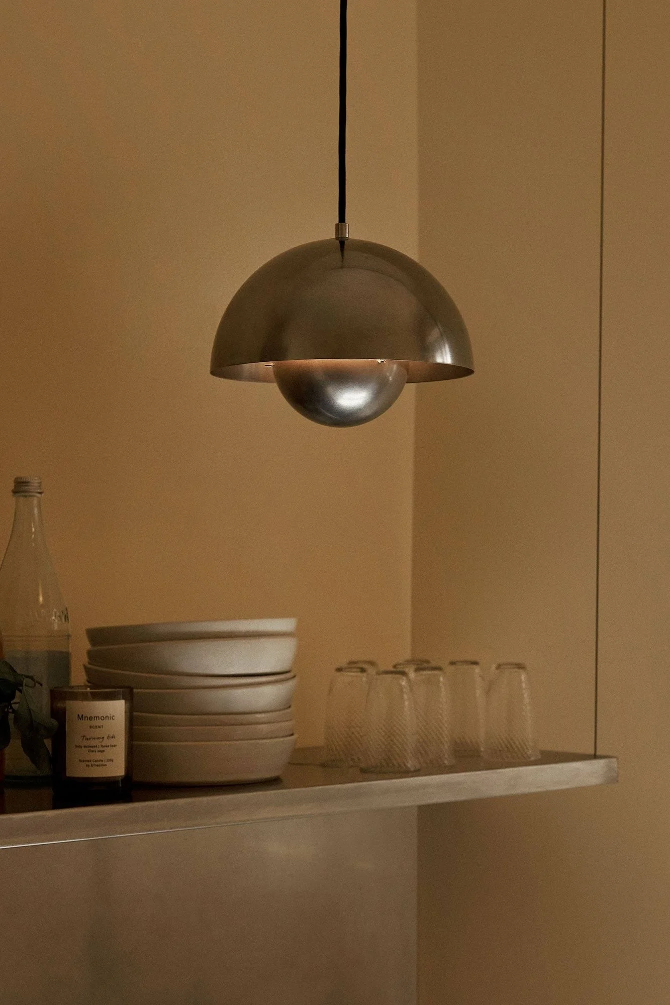 Lámpara colgante metálica de cúpula iluminando un estante con cuencos apilados, vasos, una botella y una vela.