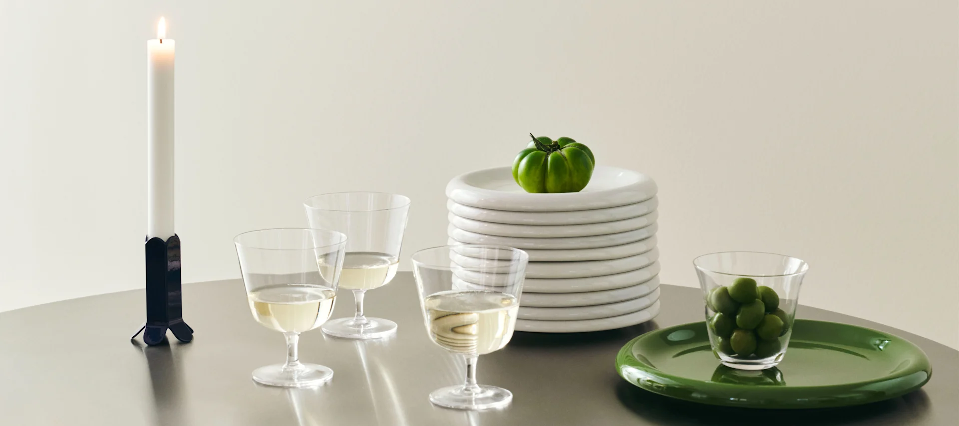 Mesa con vela, copas de vino, pila de platos blancos con tomate verde y aceitunas verdes en un plato verde.
