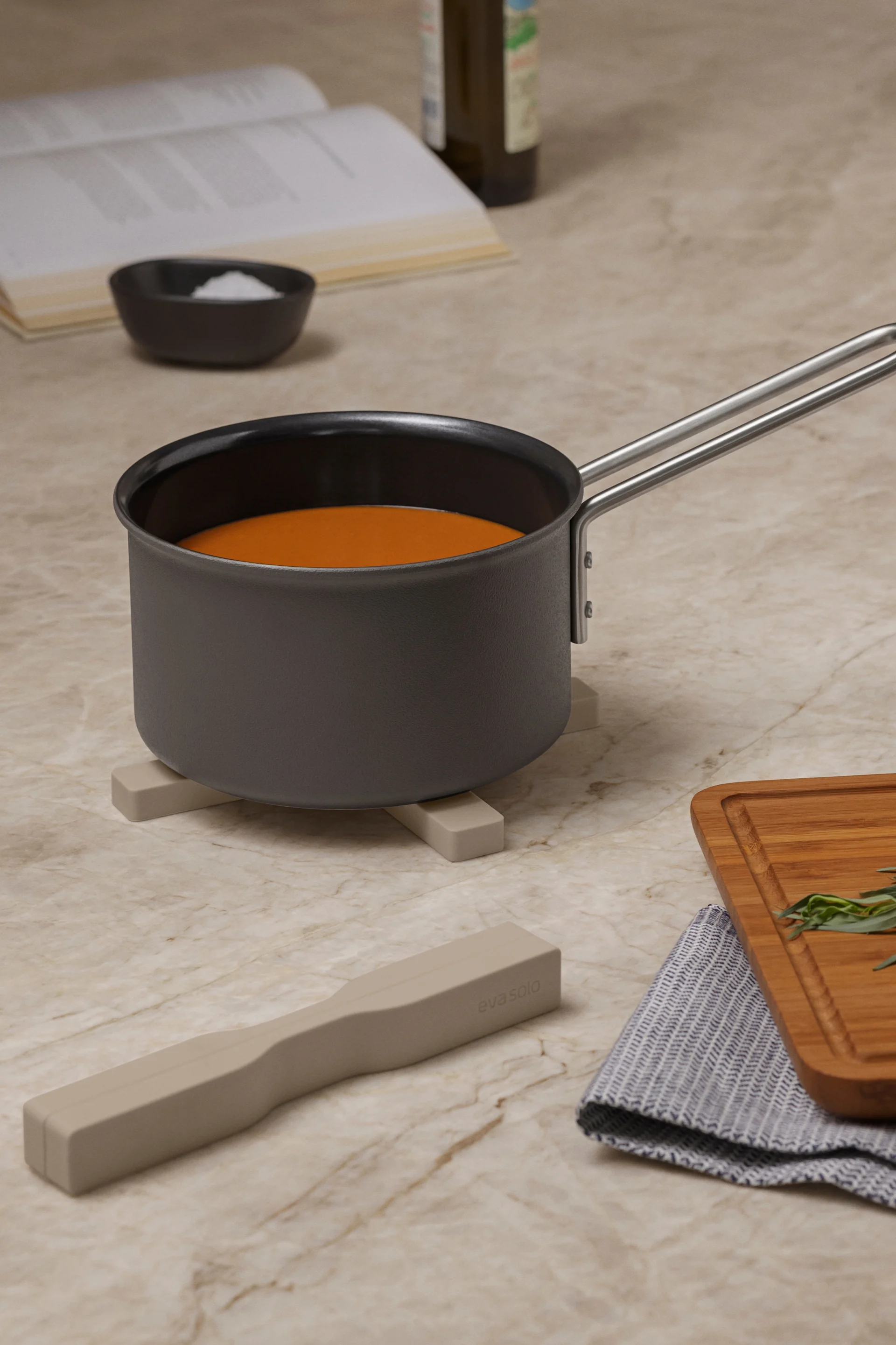 Cacerola con líquido naranja sobre un salvamanteles, con libro de cocina, sal, batidor, hierbas en tabla y utensilio en encimera de mármol.