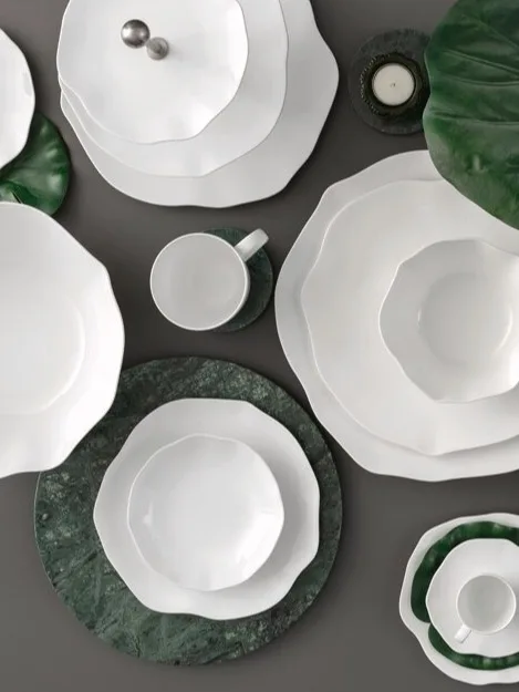 Una colección de vajilla de cerámica blanca con bordes ondulados, dispuesta con hojas verdes y posavasos de mármol sobre una superficie oscura.