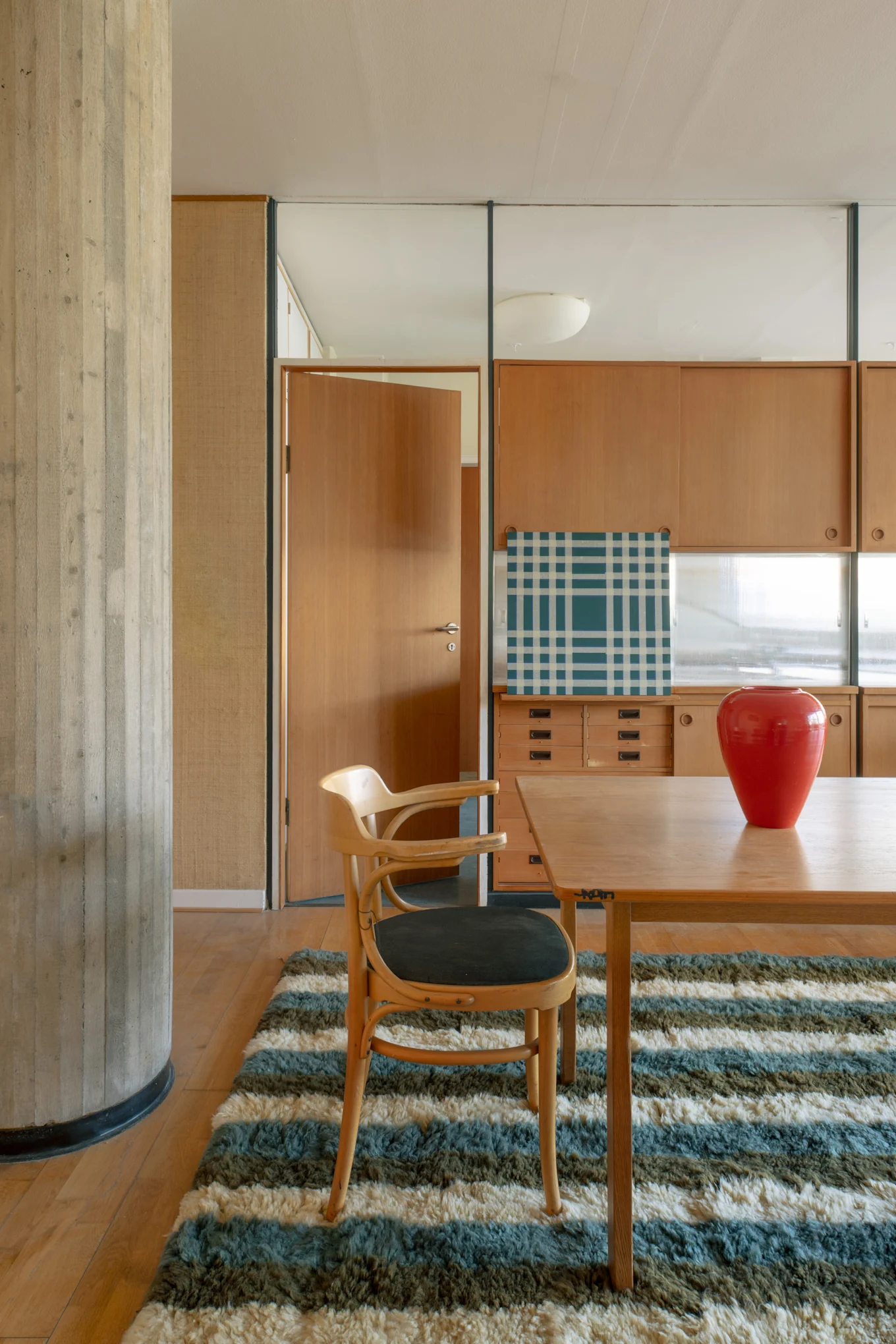 Interior moderno con mesa de comedor de madera, silla, jarrón rojo y una alfombra a rayas. Un pilar de hormigón enmarca gabinetes de madera y una puerta abierta.