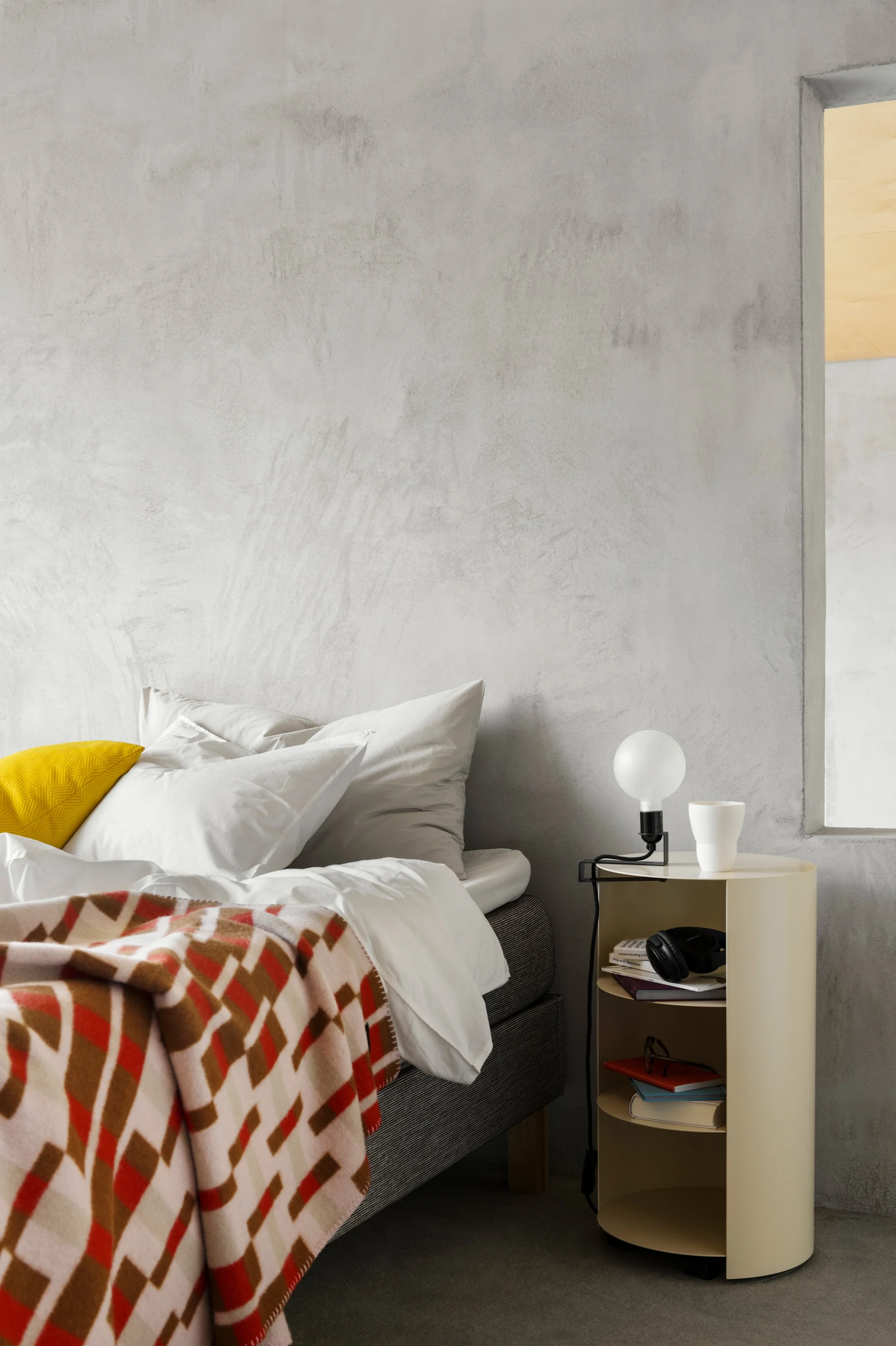 Dormitorio moderno con cama con manta estampada, almohada amarilla y mesita de noche crema con lámpara y libros.
