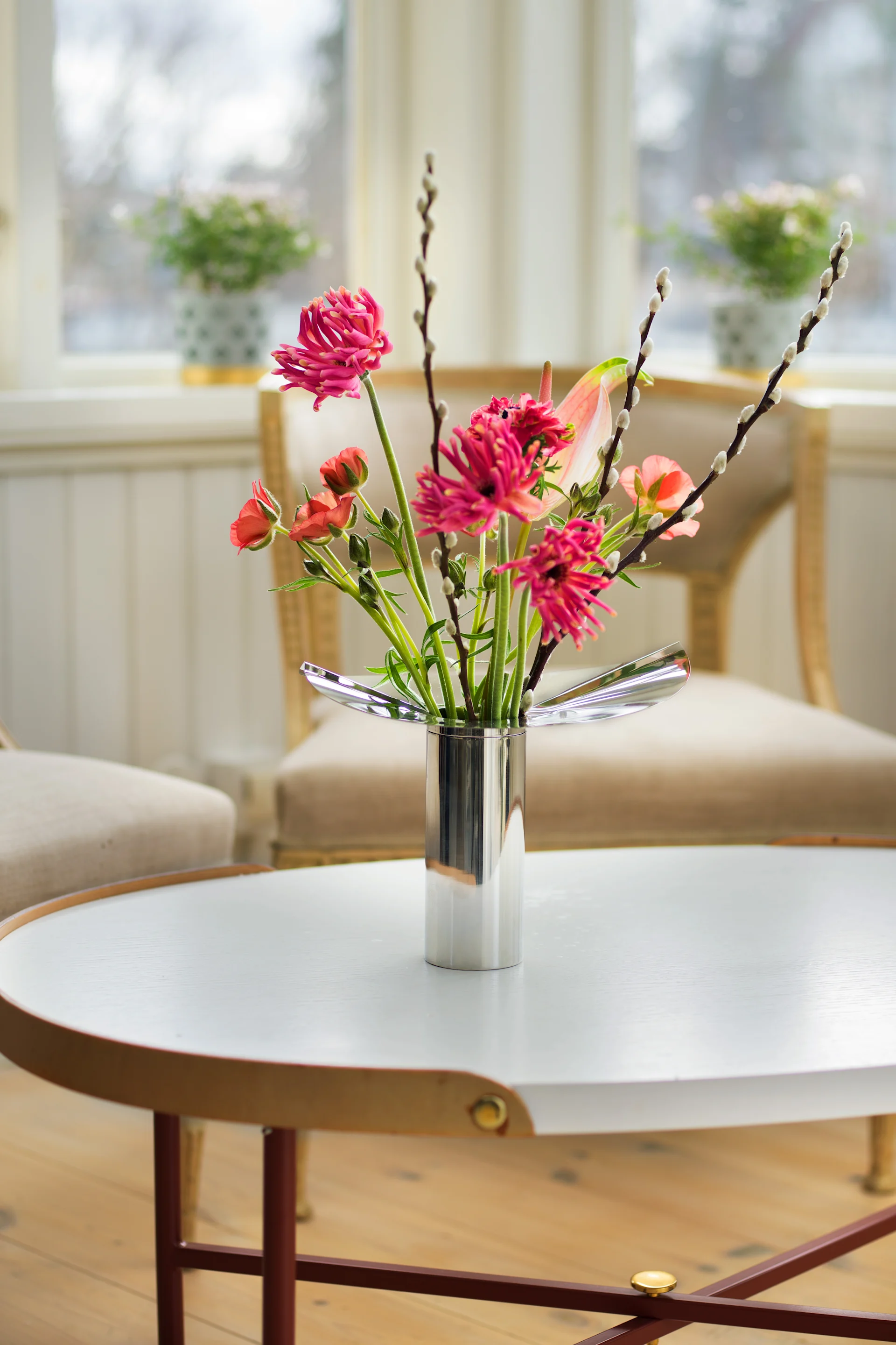 Un mesa de centro redonda está decorada con el jarrón/portavelas Löv de Klong en color cromado, con flores primaverales en su interior.