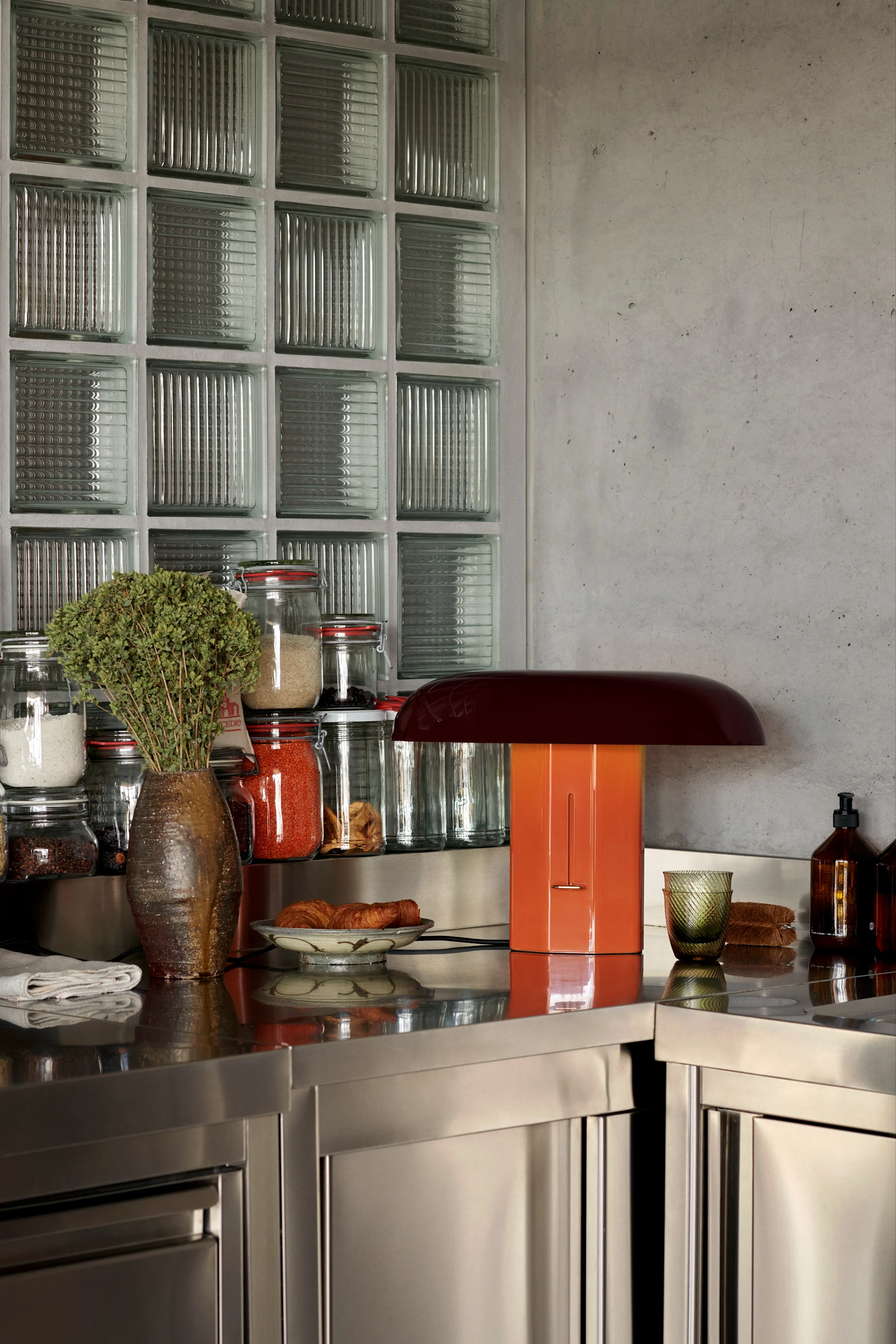 Una encimera de cocina de acero inoxidable con un jarrón marrón con hierbas, frascos de ingredientes, una lámpara naranja en forma de seta, cruasanes y botellas de vidrio.