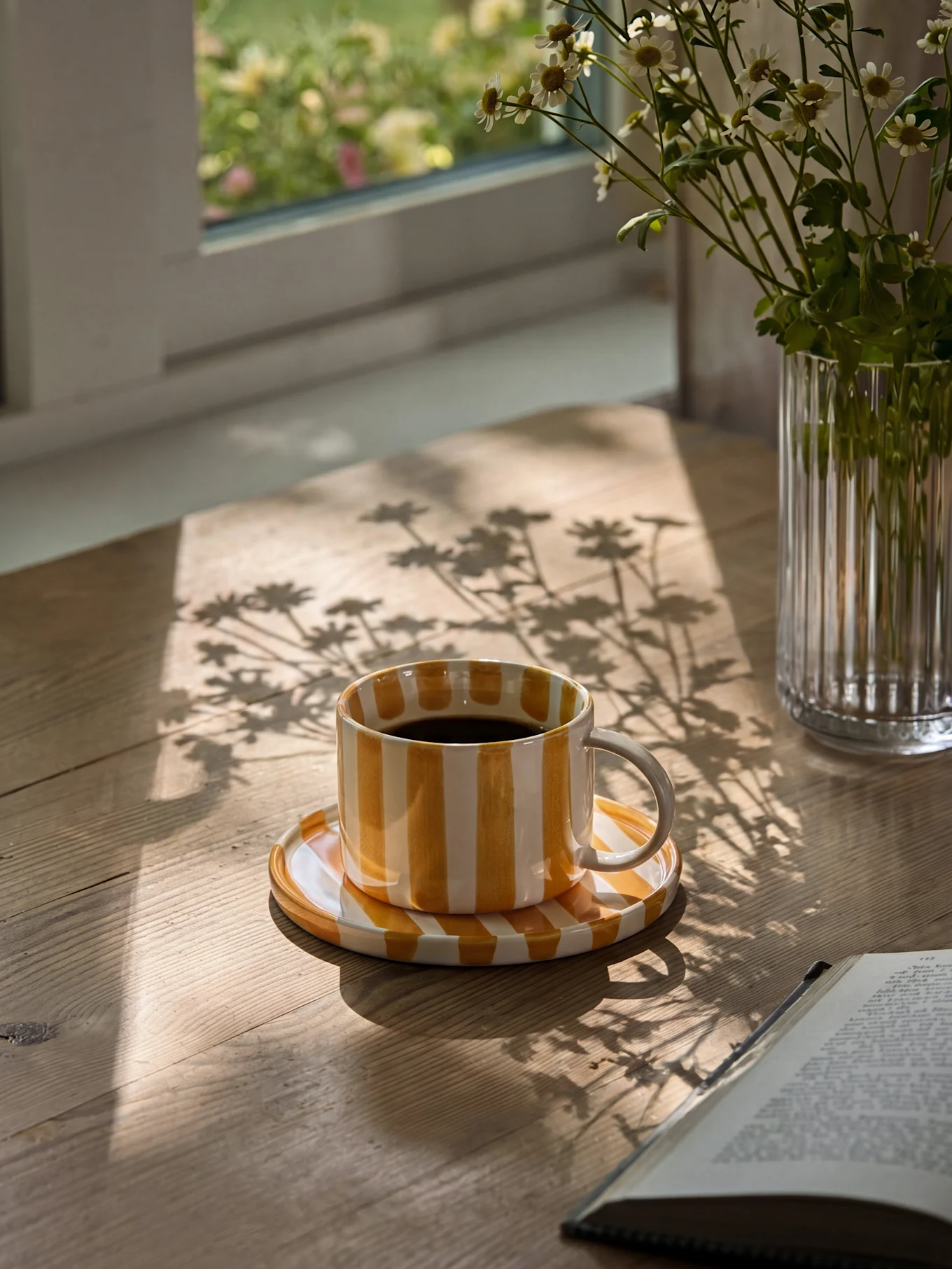 Taza de café a rayas amarillas y blancas con café negro en una mesa de madera con sombras de sol y un jarrón de margaritas.