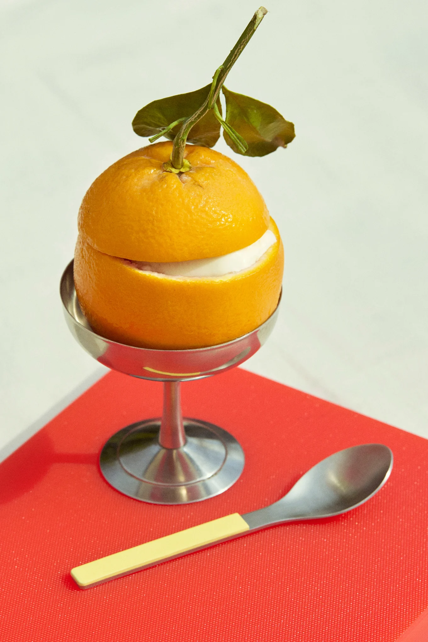 Postre de naranja con relleno blanco en copa plateada, con cuchara, sobre superficie roja.