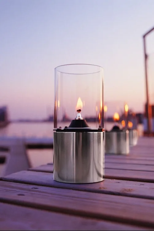 Coloca faroles y portavelas en el exterior, en el balcón, la terraza o el jardín, por ejemplo, la elegante lámpara de aceite de Stelton.