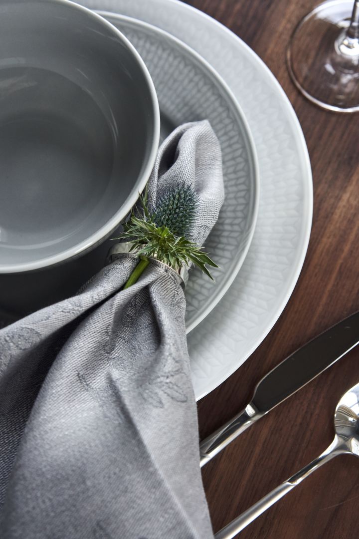 Servicio de mesa con porcelana Swedish Grace en blanco y gris y servilleta de lino en servilletero con flores como decoración. 