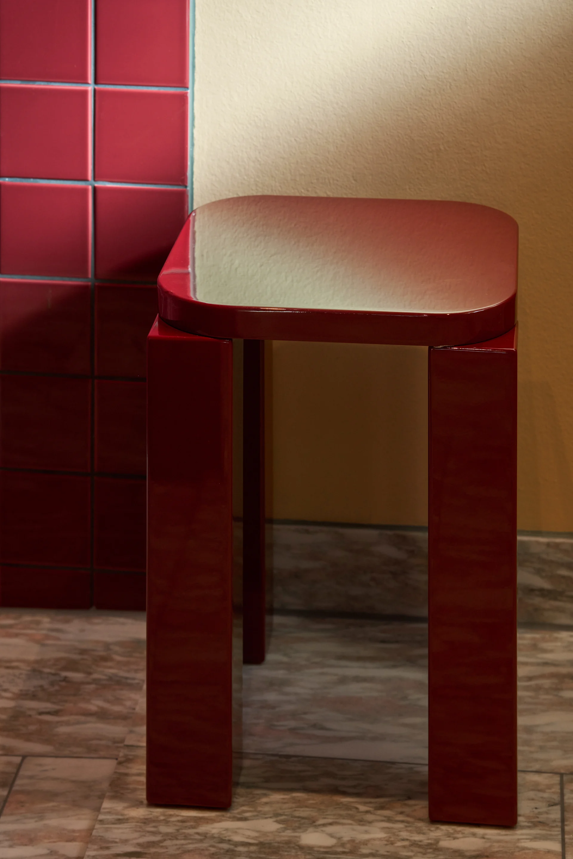 Un taburete rojo oscuro brillante con tapa rectangular y cuatro patas sobre un suelo de mármol junto a una pared de azulejos.
