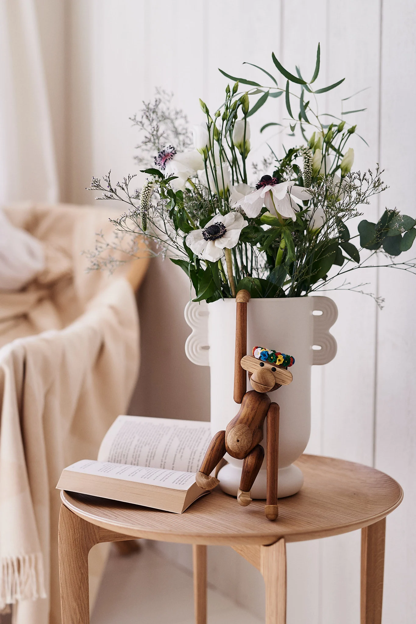 Un juguete de mono de madera cuelga de un jarrón blanco con flores blancas junto a un libro abierto en una mesa de madera.