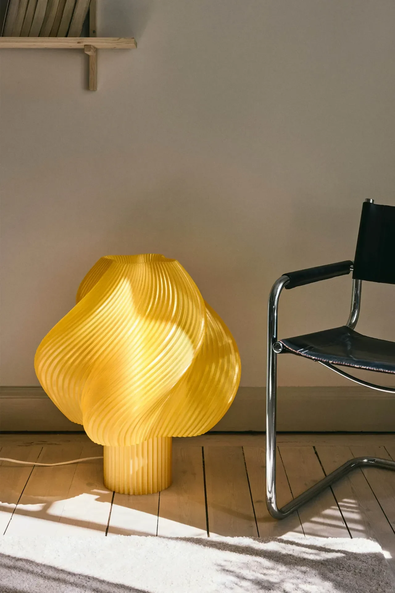 Una lámpara de mesa amarilla brillante y escultural con diseño acanalado y ondulado brilla sobre un suelo de madera junto a una silla de cuero negro y cromo.