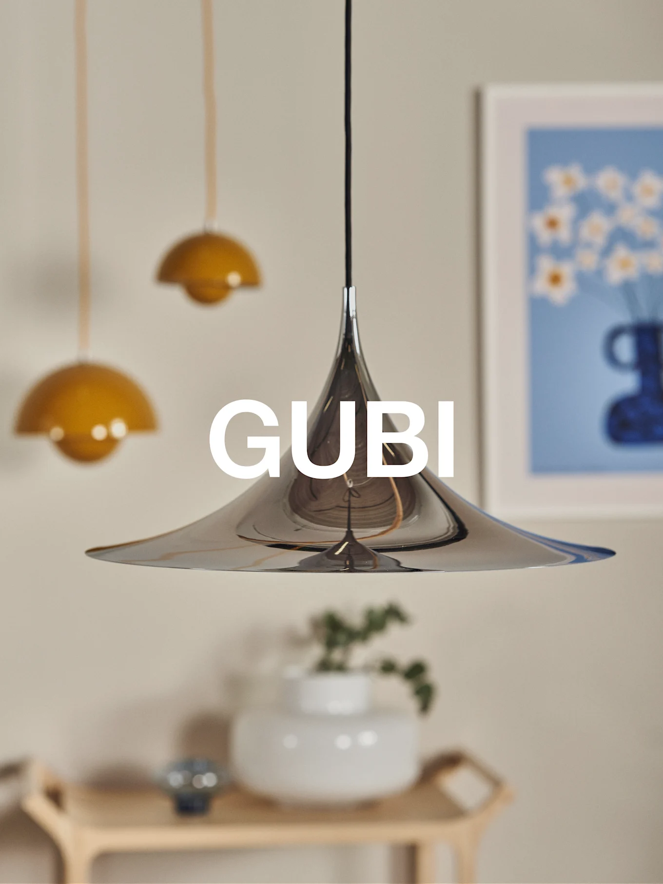Una lámpara colgante cónica de cromo pulido con el logo de GUBI, sobre un fondo borroso con otras lámparas y arte.
