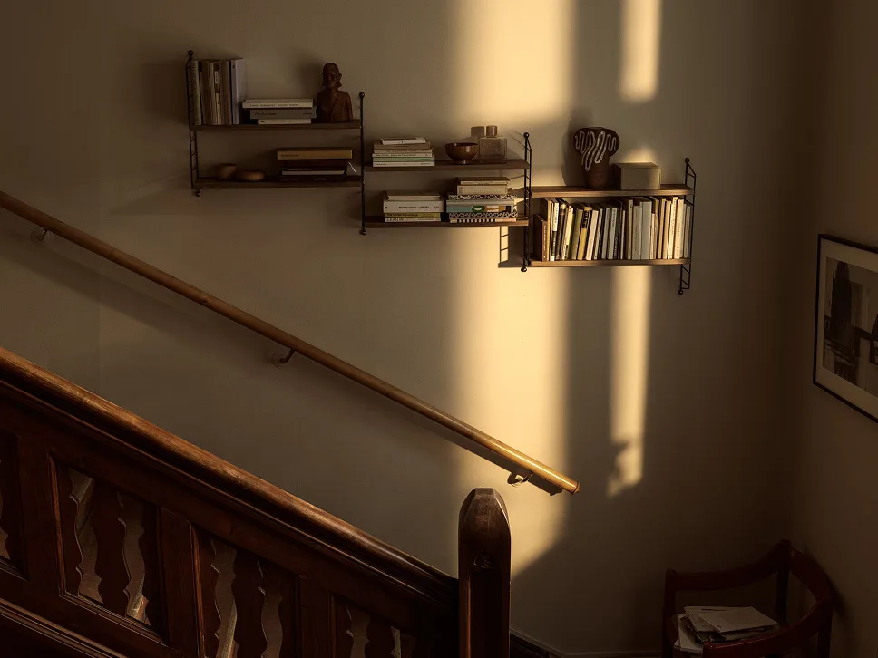 Pared soleada con estantes de madera con libros y decoración, junto a una escalera y pasamanos de madera.