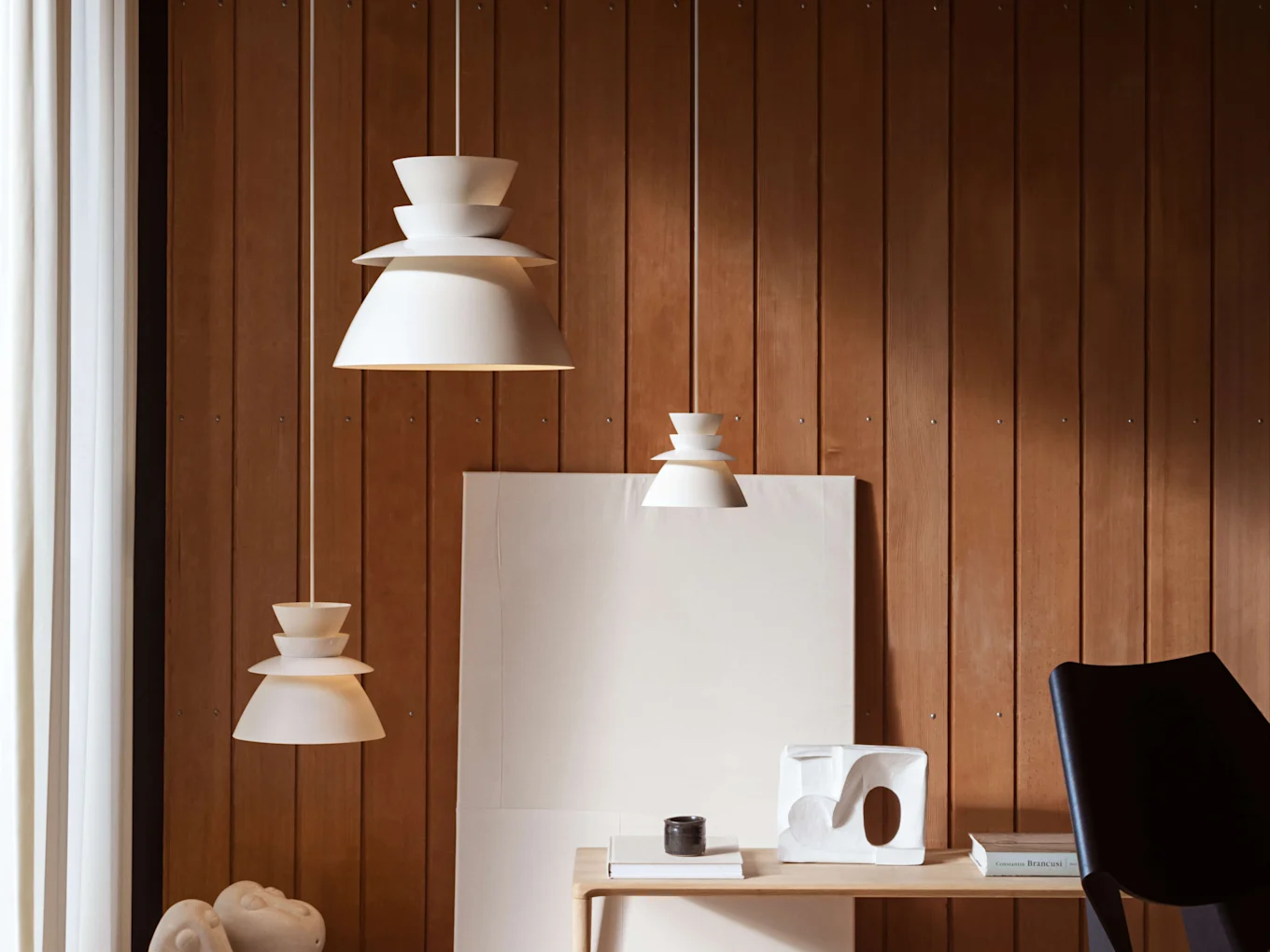 Tres lámparas colgantes blancas escalonadas cuelgan a diferentes alturas sobre una pared de madera, encima de una mesa de madera clara con decoración.