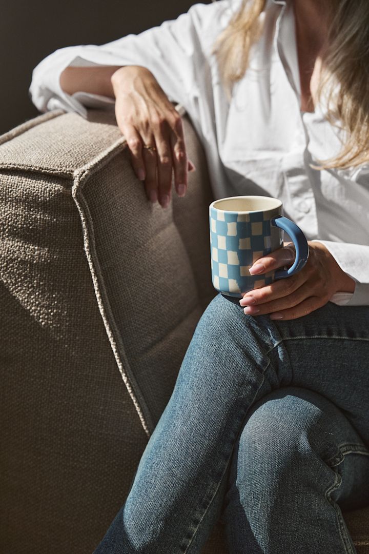 Los cuadros y las rayas son una de las grandes tendencias según las tendencias de diseño de interiores de otoño de 2024. Aquí se ve a una mujer sentada en un sillón sosteniendo una taza de cuadros azules de Byon.