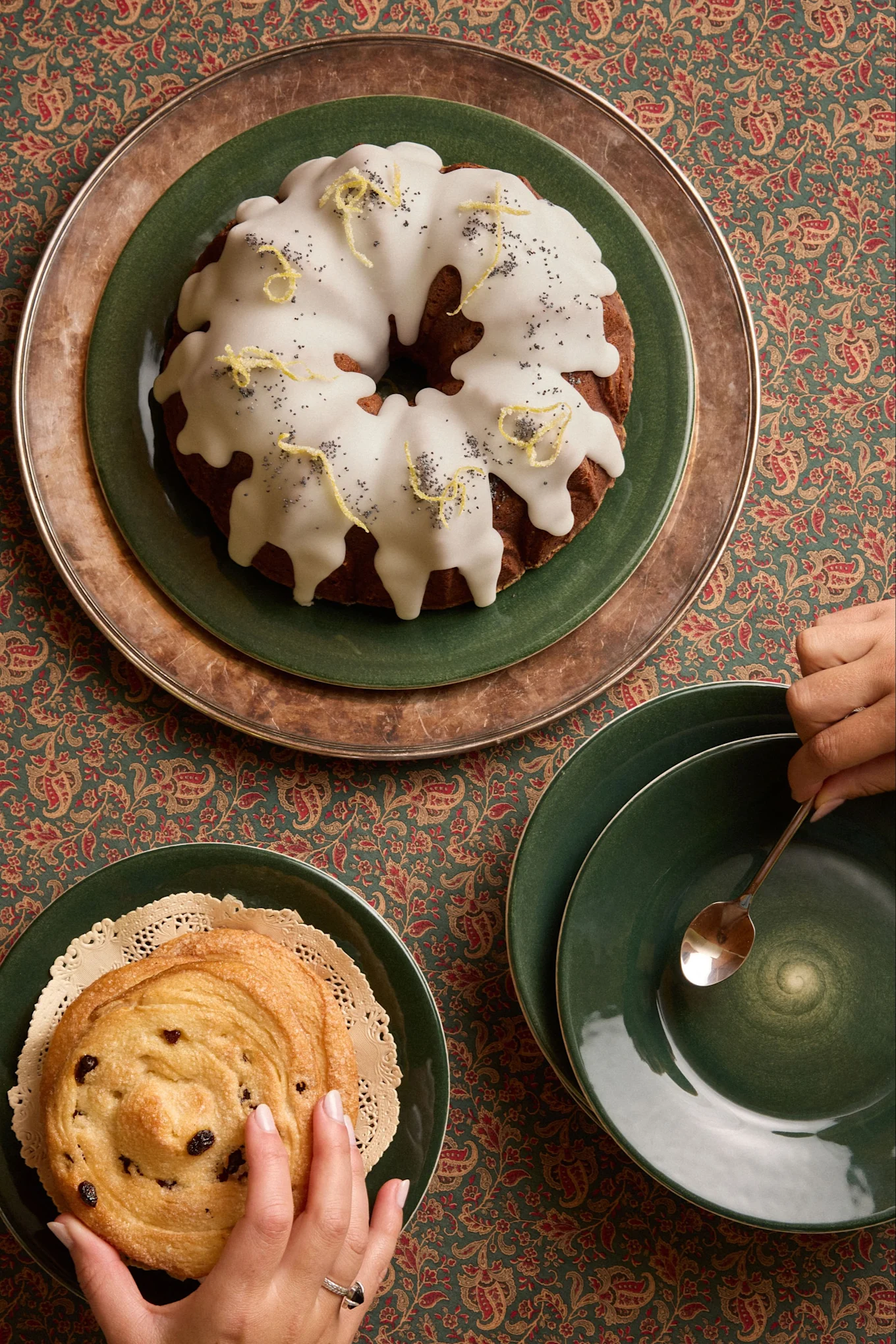 Vista superior: pastel Bundt de limón y semillas de amapola, bollo de pasas y manos sobre un mantel estampado.