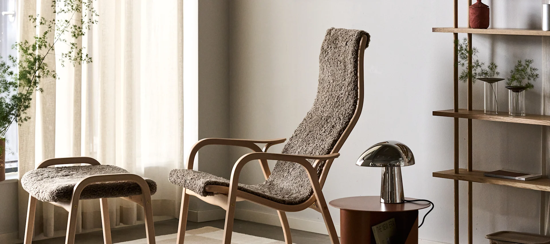 Sillón y taburete de madera clara con tapicería marrón texturizada, lámpara de mesa metálica y estantería de madera con plantas.