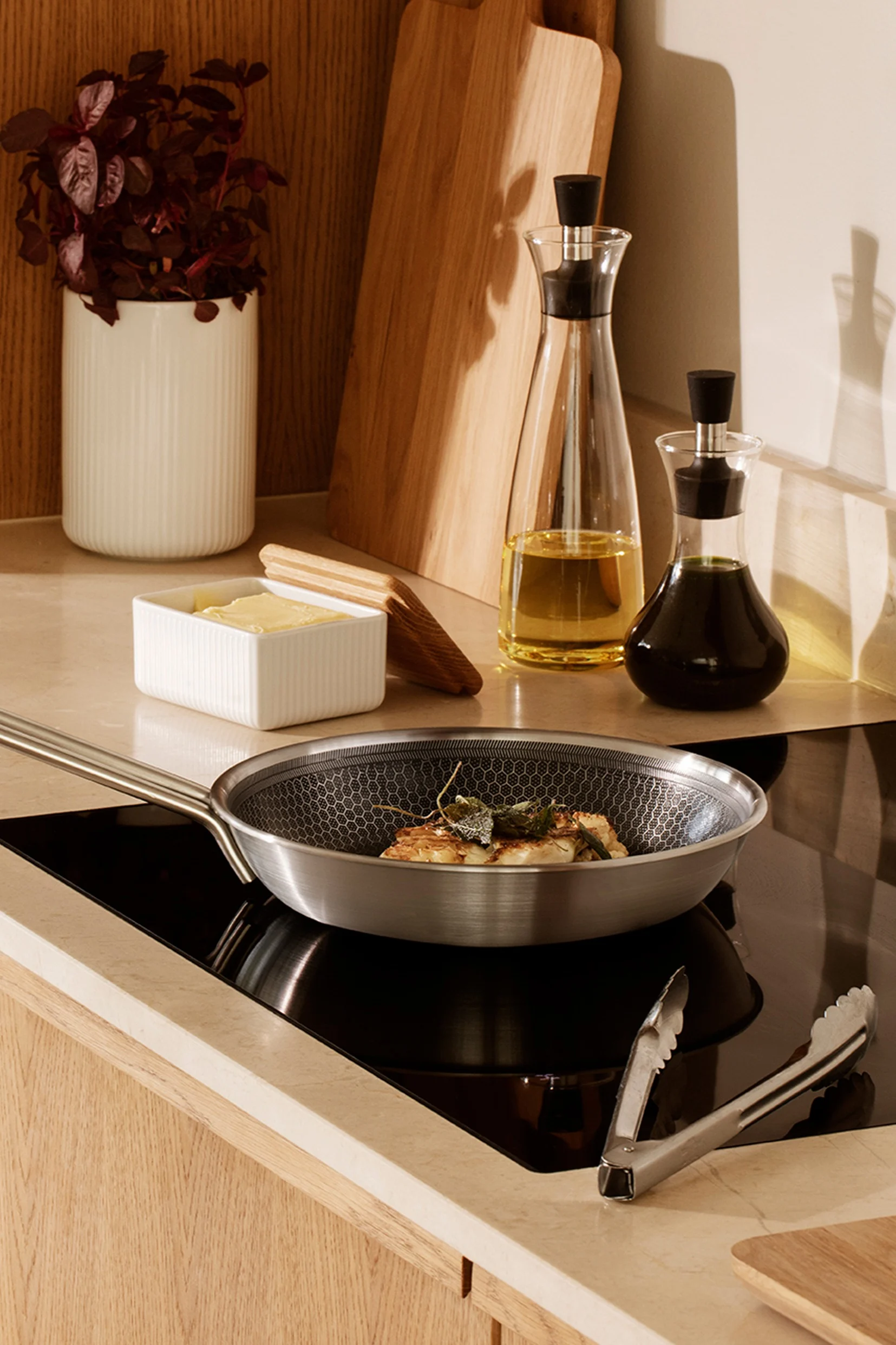 Sartén de acero inoxidable con comida en placa de inducción, junto a mantequilla, botellas de aceite y utensilios de cocina modernos.