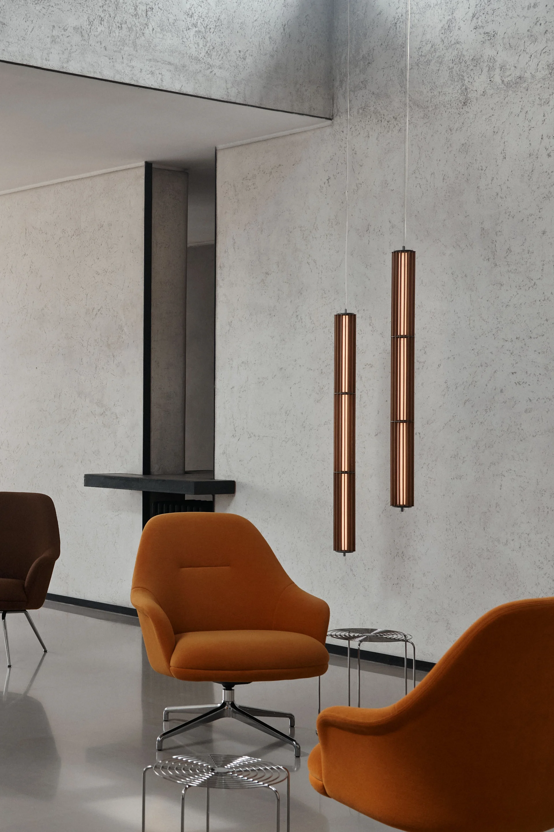 Dos lámparas colgantes cilíndricas de cobre cuelgan en una habitación moderna con sillones naranjas y paredes con textura de hormigón.